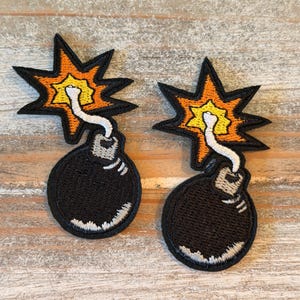May include: Two embroidered patches featuring a round black bomb with a white fuse and a bright orange and yellow star-shaped explosion. The patches have black borders and white highlights on the bottom of the bomb and the fuse.
