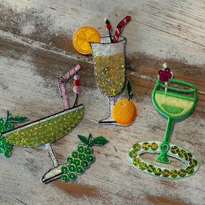 May include: Three embroidered patches shaped like cocktail glasses. One is a wide green glass with grapes, one is a tall glass with an orange slice and straw, and one is a green stemmed glass with a cherry garnish, all featuring sequins.