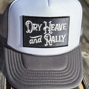 May include: A gray and white trucker hat with a black patch. The patch features the words "Dry Heave and Rally" in white script. The hat has a white front panel, a gray brim, and a black mesh back.