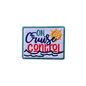 May include: Embroidered patch with the words "On Cruise Control" in blue and red script. A yellow ship's wheel is on the right, and a wavy blue line at the bottom. The patch is rectangular with a white background and a teal border.
