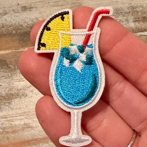 May include: An embroidered patch shaped like a cocktail glass filled with a bright blue liquid and white ice cubes. The glass is garnished with a yellow pineapple slice and features a red straw extending from the top, all outlined in white.