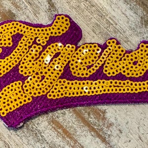 May include: Purple fabric patch with the word "Tigers" spelled out in gold sequins.