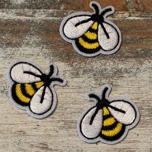 May include: Three embroidered bee patches. Each patch features a white body, black stripes, and a yellow abdomen. The bees have black antennae and are outlined in gray. The patches are on a weathered, light-colored wooden surface.