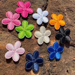 May include: Ten embroidered iron-on patches in various colors, each featuring a five-petal flower design. The patches are arranged in a circle on a brown, textured surface.