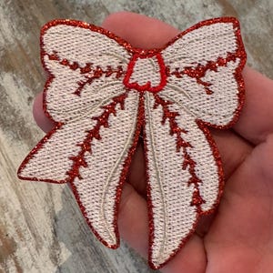 May include: A white and red embroidered bow applique. The bow features a detailed design with red glitter accents outlining the edges and center. The bow is held in a hand.