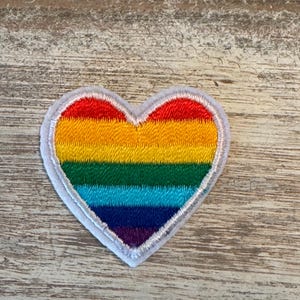 May include: A heart-shaped patch with a rainbow design. The patch features horizontal stripes in red, orange, yellow, green, blue, and purple. The patch is outlined with a white border and a gray stitched edge.