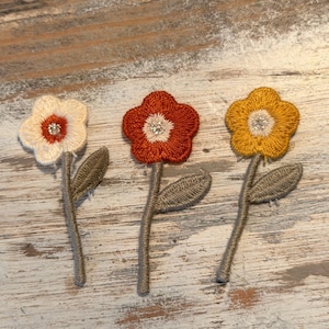 May include: Three embroidered flower patches in cream, rust, and gold. Each flower has a central rhinestone and is attached to a stem with a leaf. The patches are likely for embellishing clothing or crafts.