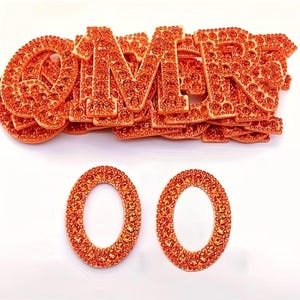 May include: Orange rhinestone letters spelling out the word "OMMR" and two additional "O" letters. The letters are bedazzled with small, orange rhinestones. The letters are stacked on top of each other, with the two "O" letters below.