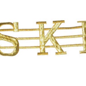 May include: Gold embroidered letters spelling "SKI" on a musical staff. The letters are in a serif font and are stitched with a metallic gold thread. The musical staff has three horizontal lines.