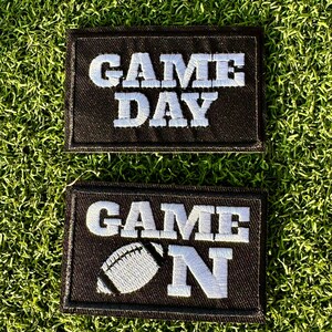 May include: Two black fabric patches with white embroidered text. The top patch says "GAME DAY". The bottom patch says "GAME ON" with a white football graphic.