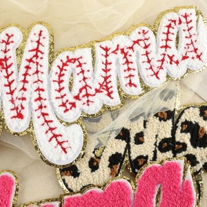May include: White felt letters spelling "Mama" with red stitching and gold glitter trim. The letters are layered on top of a beige tulle fabric.