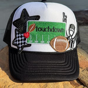 May include: A black and white trucker hat with a white front panel. The hat features embroidered patches of a football, a football field with the word "touchdown", stars, and a referee. The hat has a black brim and mesh back.