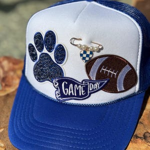 May include: A blue and white trucker hat with a blue brim. The hat features a blue paw print, a football, and a banner that says "Game Day". A gold safety pin with pearls and a heart charm is also attached.