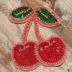 May include: A beaded cherry applique with red sequined cherries and green sequined leaves. The applique is outlined with pink beads and thread, creating a dimensional effect. The design is a playful and decorative embellishment.