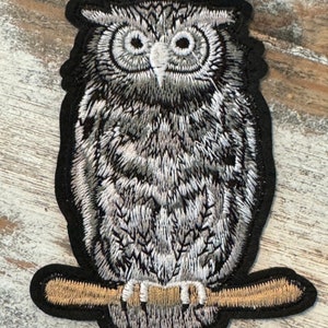May include: A gray and white embroidered owl patch with a brown branch. The owl is perched on the branch with its wings folded. The patch is on a wooden background.