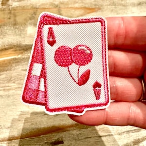 May include: Embroidered patch featuring a playing card design. The card is white with a red border and depicts the Ace of Hearts with a pair of cherries. The patch is held in a hand.