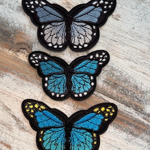 May include: Three embroidered butterfly patches in shades of blue, black, and yellow. The butterflies have detailed wing patterns and are set against a light-colored wooden surface. The patches are likely for clothing or craft projects.