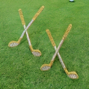 May include: Two pairs of gold golf clubs crossed over each other on a green putting green. A white golf ball is in the upper right corner of the image.