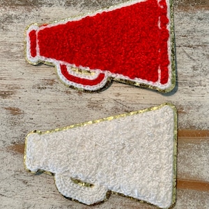 May include: Two felt megaphones, one red and one white, with gold glitter trim. The megaphones are on a wooden background.