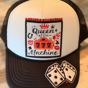 May include: A white and black trucker hat featuring a rectangular patch with the text Queen of the Machine and a red 777 slot machine graphic. The patch is accented with red rhinestones along the top edge, and a pair of dice is embroidered on the brim.