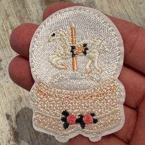 May include: White embroidered iron-on patch of a carousel horse inside a snow globe with pink flowers and a white base.