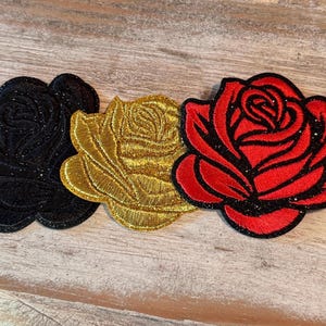 May include: Three embroidered rose patches featuring a black rose, a gold rose, and a red rose. Each patch has a distinct floral shape with detailed stitching and a subtle glittery finish on the petals, arranged side by side in a horizontal row.