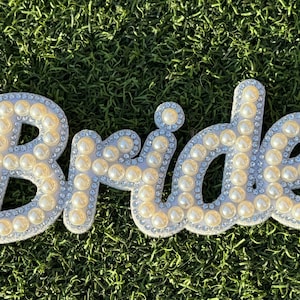 May include: White "Bride" sign decorated with pearl-like beads and rhinestones.