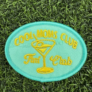 May include: A turquoise blue oval-shaped patch with yellow embroidered text that reads "Cool Moms Club" and a martini glass with a yellow outline. The text "Fun Club" is also embroidered in yellow below the martini glass.