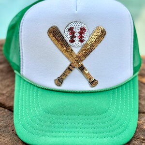 May include: A green and white trucker hat featuring a sequined patch on the front panel. The patch displays two crossed gold sequin baseball bats behind a white sequin baseball with red sequin stitching. The hat has a green mesh back and brim.