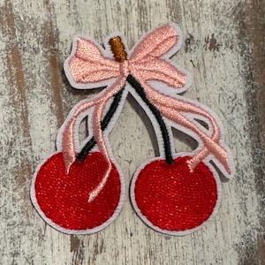 May include: Two red cherries with green stems and a pink bow. The cherries are embroidered on a white fabric.