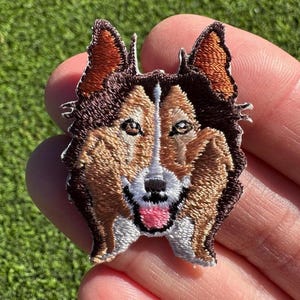 May include: Embroidered patch of a brown and white dog with a pink tongue sticking out. The dog has brown eyes and a white patch on its chest.
