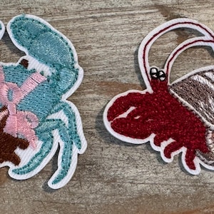May include: Two embroidered patches featuring sea creatures. One is a blue crab with a brown bear and pink ribbon. The other is a red crab carrying a shell. Both patches have white borders.