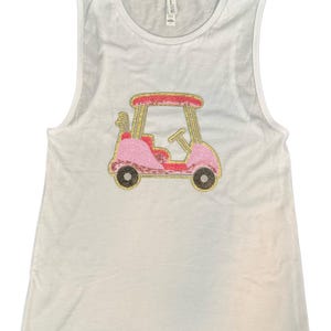 May include: White sleeveless shirt with a pink and gold sequined golf cart design. The golf cart features a pink body, gold accents, and black wheels. The shirt is made of a soft, lightweight material.