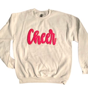 May include: White sweatshirt with the word "Cheer" in pink glitter lettering.