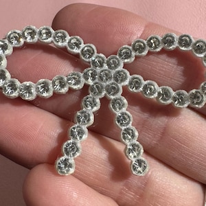 May include: A sparkling bow-shaped brooch. The brooch is made of clear, round rhinestones set in a white backing. The bow design is symmetrical, with two loops and two streamers. The brooch is held in a hand.