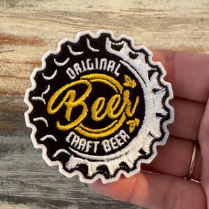 May include: Embroidered patch in the shape of a beer bottle cap. The patch features the words "ORIGINAL Beer CRAFT BEER" in yellow and white stitching on a black and white background. The patch is circular with a scalloped edge.