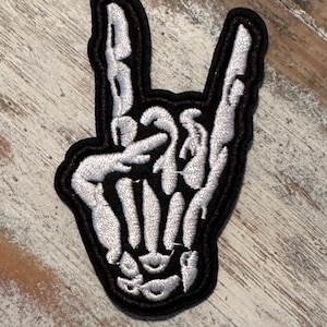 May include: Embroidered patch featuring a white skeleton hand making the rock and roll hand gesture, outlined in black. The patch is designed to be ironed on or sewn onto clothing or accessories.