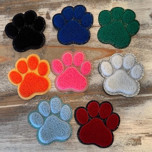 May include: A collection of seven colorful paw print patches. The patches are in various colors, including black, blue, green, orange, pink, white, and red. Each patch has a glittery border.