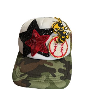 May include: A white baseball cap with a camouflage brim. The cap has a red and black star, a baseball, and a yellow and black bee patch.