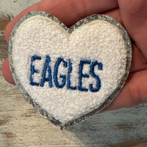 May include: A heart-shaped patch with the word "EAGLES" embroidered in blue on a white, textured background. The patch has a silver glitter border and is held in a hand.