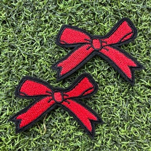 May include: Two red embroidered bow patches with black outlines are displayed against a green, textured background. The bows have a classic shape, with detailed stitching. These patches could be used for clothing or craft projects.