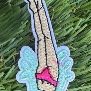 May include: Embroidered patch featuring a stylized illustration of a raised fist with a light skin tone. The fist is positioned above a pair of legs wearing pink underwear, all set against a light blue and white background.