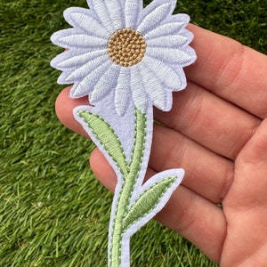 May include: White felt daisy flower with a brown center and green stem and leaves. The flower is embroidered with white thread and the stem and leaves are embroidered with green thread.