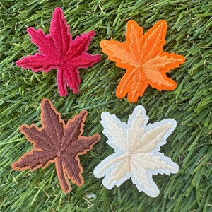 May include: Four embroidered felt maple leaf patches in different colors: red, orange, brown, and cream. The patches are on a green artificial grass background.