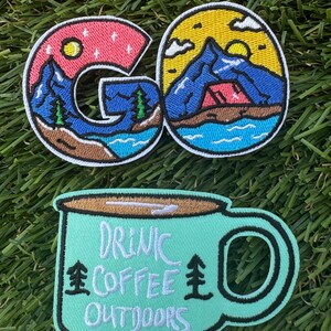 May include: Two embroidered patches. The first patch is a colorful depiction of a mountain scene with a tent and the word "GO". The second patch is a green mug with the text "DRINK COFFEE OUTDOORS".