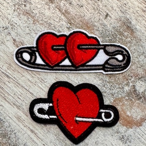 May include: Two red embroidered heart patches with a safety pin going through them. The top patch has two hearts and the bottom patch has one heart.