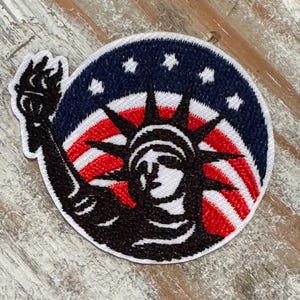 Pu&ograve; includere: Toppa ricamata con la Statua della Libert&agrave; in nero, in piedi davanti a una bandiera americana con strisce rosse e bianche e stelle blu.