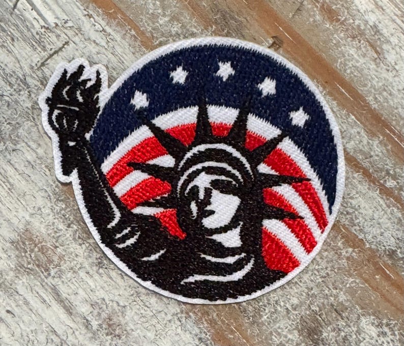 Pu&ograve; includere: Toppa ricamata con la Statua della Libert&agrave; in nero, in piedi davanti a una bandiera americana con strisce rosse e bianche e stelle blu.