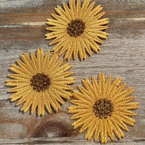 May include: Three embroidered sunflower patches with yellow petals and brown centers are arranged on a wooden surface. The patches are detailed with textured stitching, showcasing a craft or DIY theme.