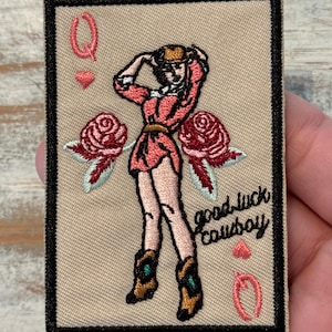 May include: Embroidered patch featuring a cowgirl on a playing card. The card has a black border and the text "good luck cowboy". The cowgirl is wearing a pink dress and cowboy boots. Red roses are on either side of the cowgirl.